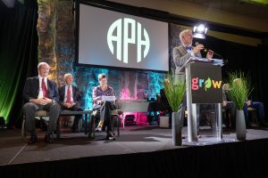 APH's president stands behind a podium reading "grow" on a stage in front of a large audience. 