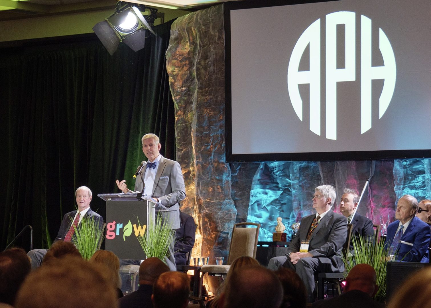 Craig Meador stands on stage behind a podium that reads 'grow' in front of a large screen with the APH logo