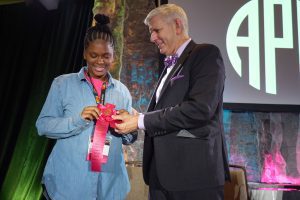 A girl stands to the left receiving an award for her art from APH's President, standing on the right. 