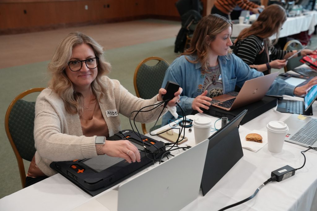 A woman sits at a desk where she is working on pugging in her new Monarch, she smiles up at the camera while still holding the cord in the air.