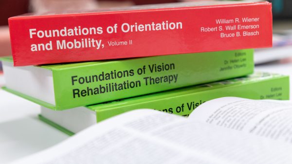 The red hardback Foundations of O&M volume II on top of a stack of green Foundations books. The title and editors' names are printed on the spine in white text.