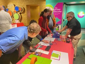 Four people stand around a red table. On it are APH STEM products like snap circuits and code and go mouse