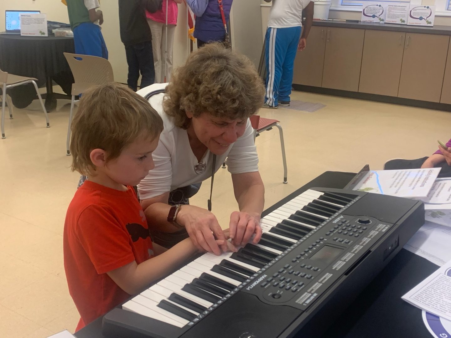 A woman stands in front a child, placing his hands on the keys to help him play.