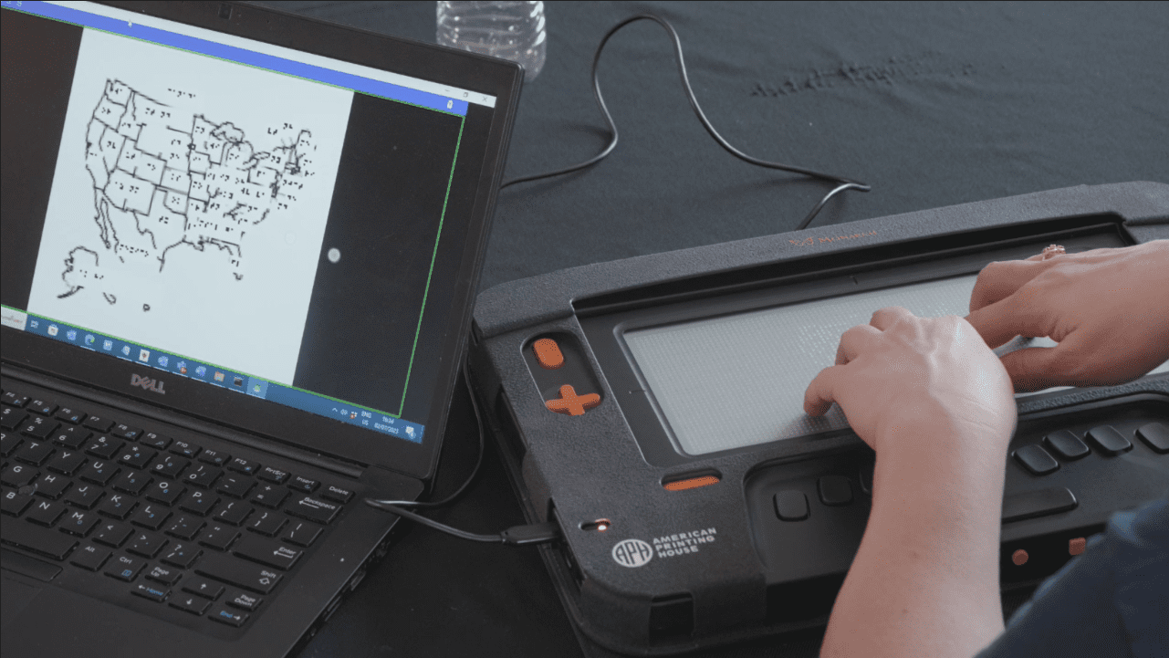 A screenshot of a woman’s hands exploring a tactile graphic of the U.S. map while it’s displayed on a laptop screen next to her Monarch.