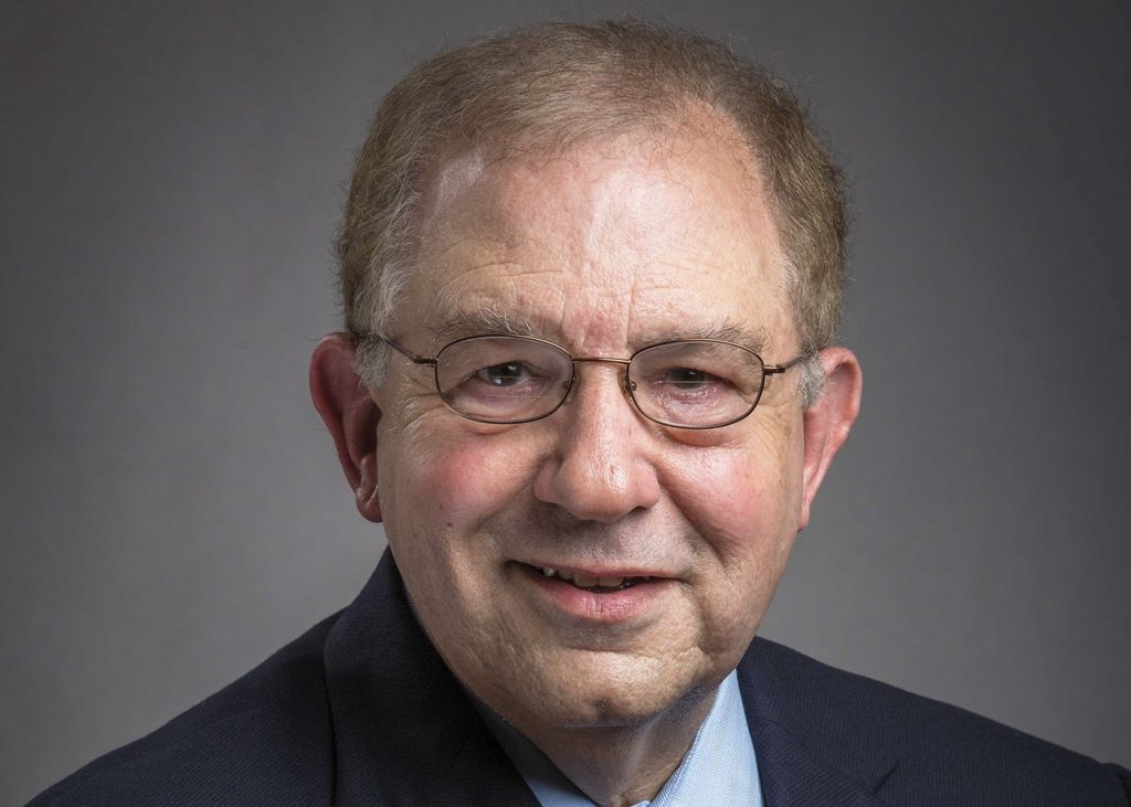 An older man with glasses smiles wearing a black suit in front of a grey background.