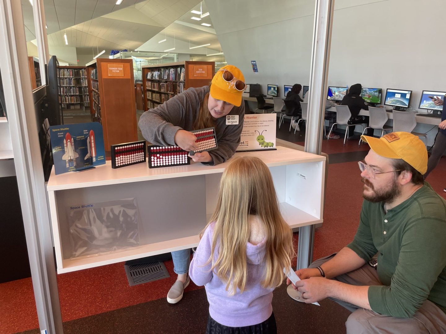 A woman wearing a gold APH hat shows a young girl and a man how to use an abacus.