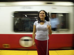 A woman stands, holding her cane, in front of a train passing by.