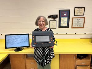 Miriam stands in front of a yellow desk while holding her Monarch. The display on the Monarch is also being shown on a computer screen next to her.