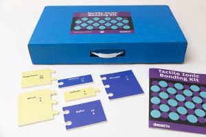 Yellow and blue kit pieces lay on a white table in front of a blue box and beside a blue and purple instruction book.