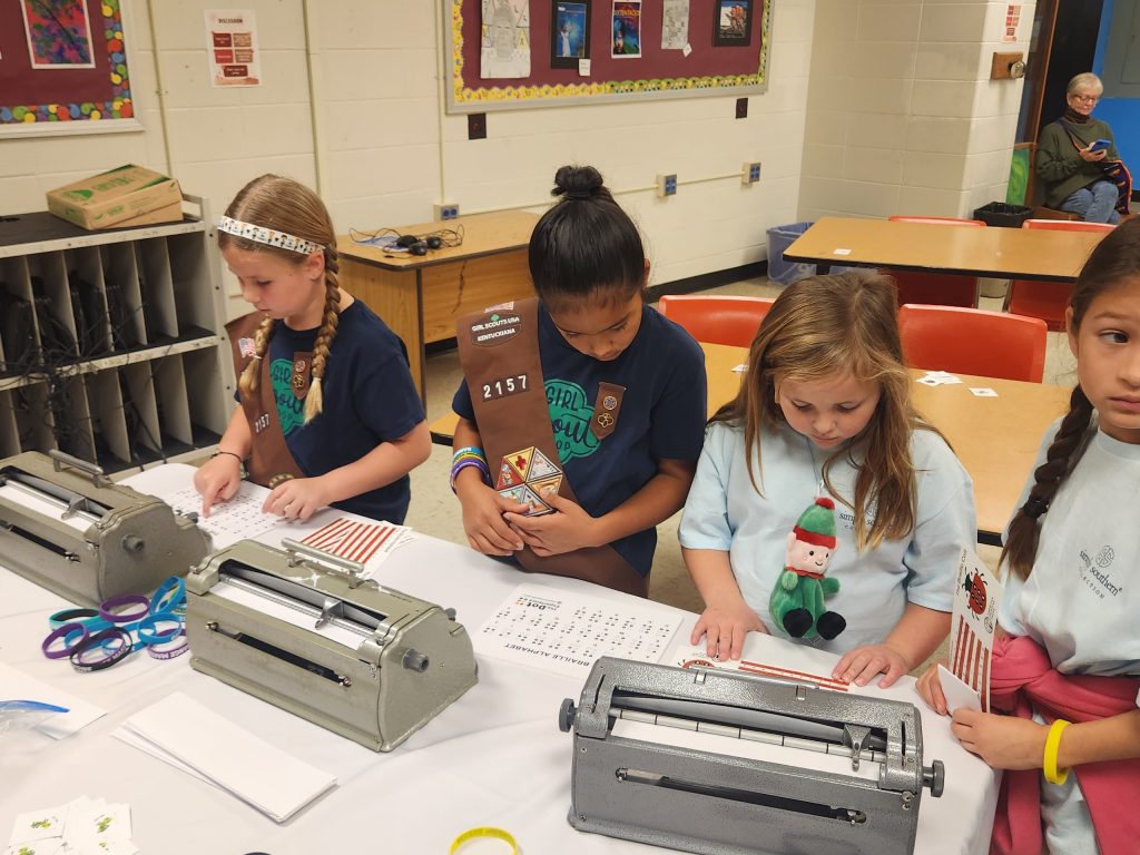 Four girls with girl scout uniforms stand around braille typewriters and braille alphabet sheets.