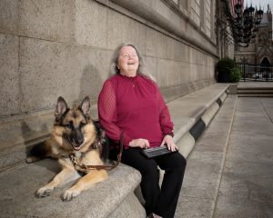 Judy smiles broadly as she holds a braille display while sitting next to her guide dog.