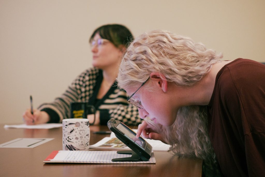 A girl wearing glasses leans down to view her screen readers as peers take notes behind her.