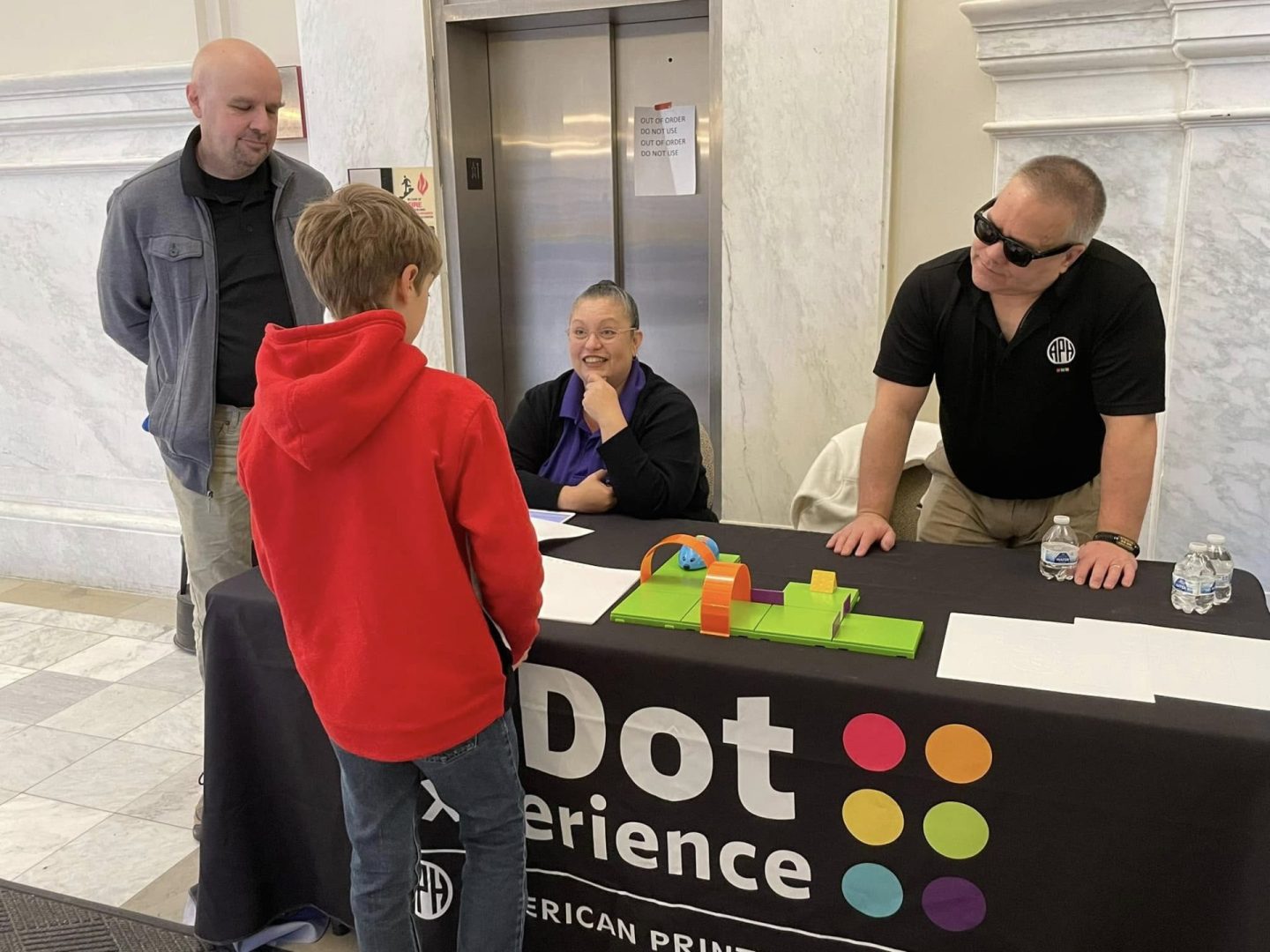 A boy stands in front of a table that reads "the Dot Experience" while talking to two APH employees sitting on the other side of the table.