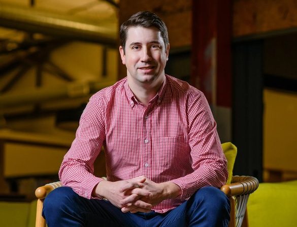 Photo of Chris Moizer. He is sitting in a chair with his hands together. He is wearing a red shirt, blue jeans, and brown shoes.