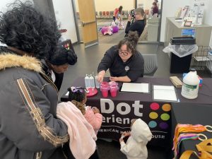 A Dot Experience volunteer helps two children and two adults make slime at a table with The Dot Experience logo on it.