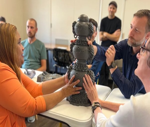 A group of people touch a statue. They are seated at a table with their hands outstretched.