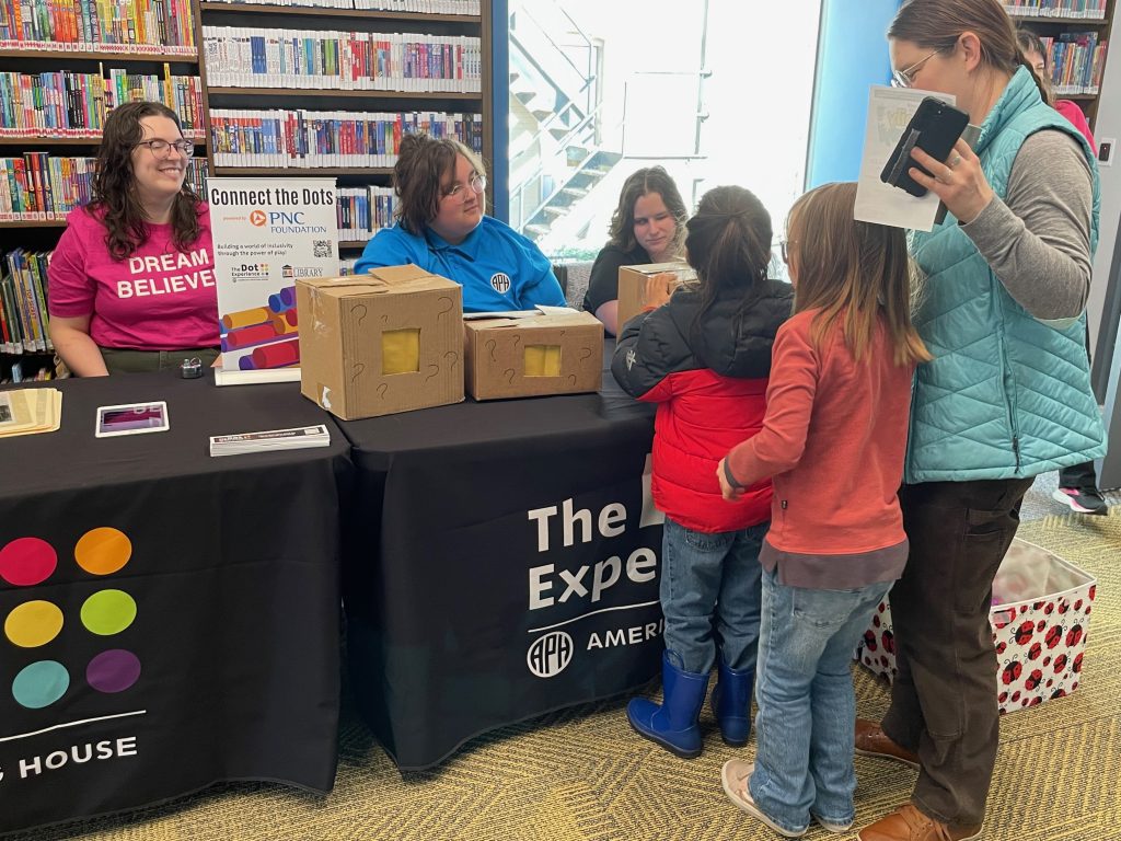 Three people sit behind a table as a woman and two children examine cardboard boxes with question marks on them that are sitting on the table. A sign with the words “Connect the Dots” on it is also visible on the table.