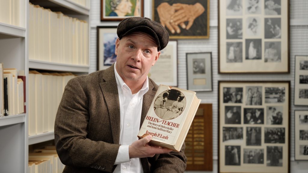 Justin Gardner sits in front of shelves filled with tan boxes. He is holding a book with a photo of Helen Keller on the cover.